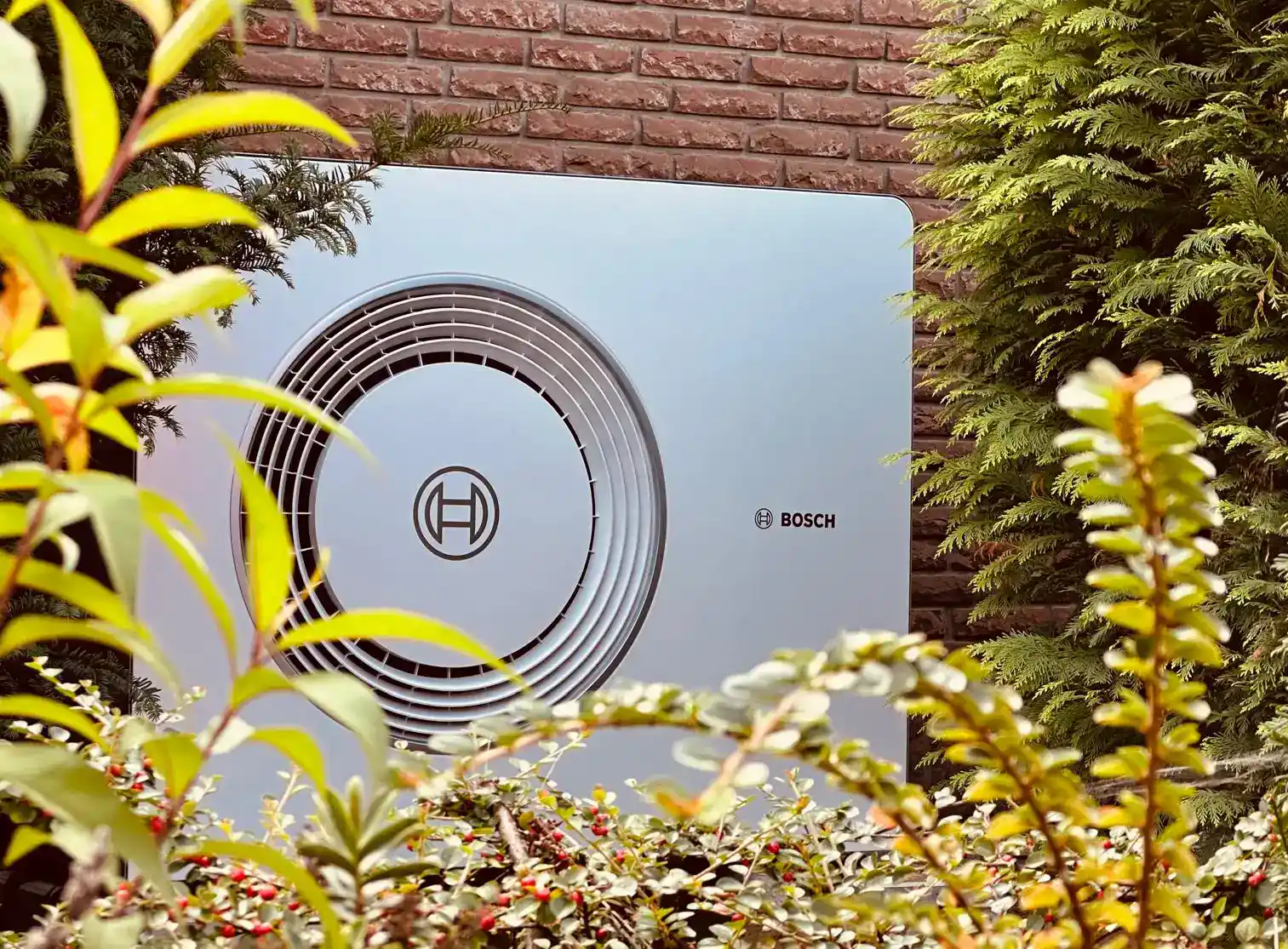 Außengerät einer Bosch Wärmepumpe neben einer Hauswand, umgeben von Gartenpflanzen